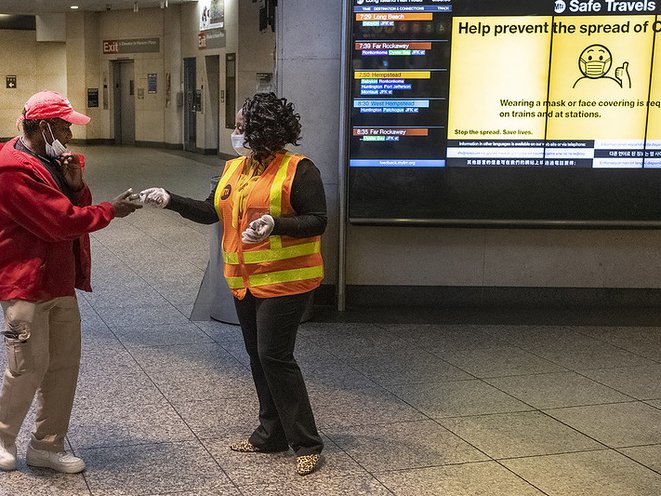 A NYC subway worker wearing a surgical mask hands out a bottle of hand sanitizer to a commuter.