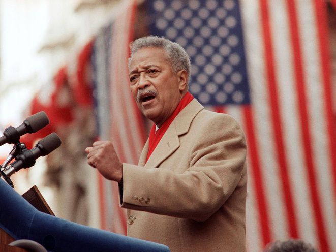 David Dinkins, in a tan coat and red scarf, stands at a lectern and in front of  American flags