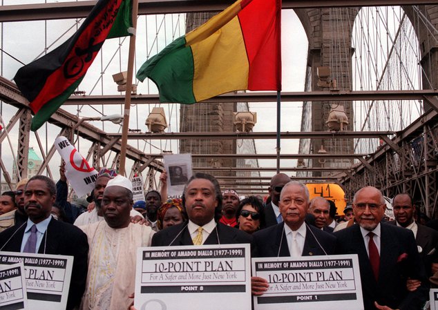On the Brooklyn Bridge, numerous marchers hold flags in Red, Gold, and Green; in the front are the Saikou Diallo, Rev. Al Sharpton, former Mayor Dinkins, and PercySutton, who are holding signs "In Memory of Amadou Diallo"