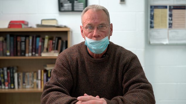 A photo of prisoner Bobby Ehrenberg, wearing a mask, sitting in front of a shelf of books.