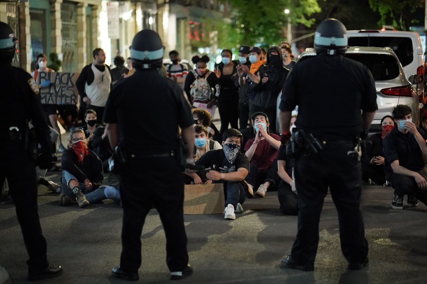 Protesters sit on the street with riot officers, with their backs turned to the camera, facing them