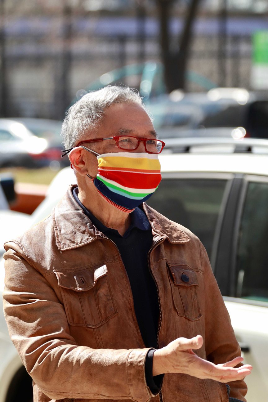 Rocky Chin, wearing  glasses and a colorful striped mask, stands on a sidewalk