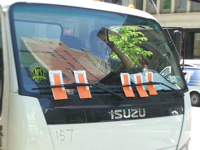 The front windshield of a white truck with several red parking tickets under the windshield wipers.