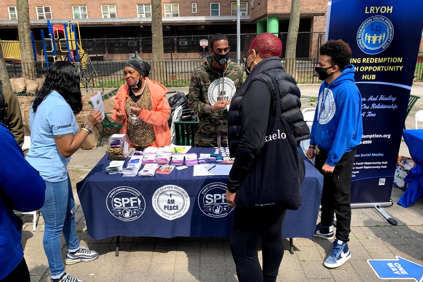 Members of Street Corner Resources stand at a table, which has a covering with the group's name, and they hand out pamphlets to people