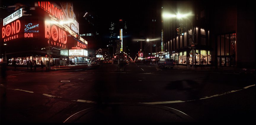 A color photograph of Times Square at night, with the red lights of the Bond's sign visible on the left