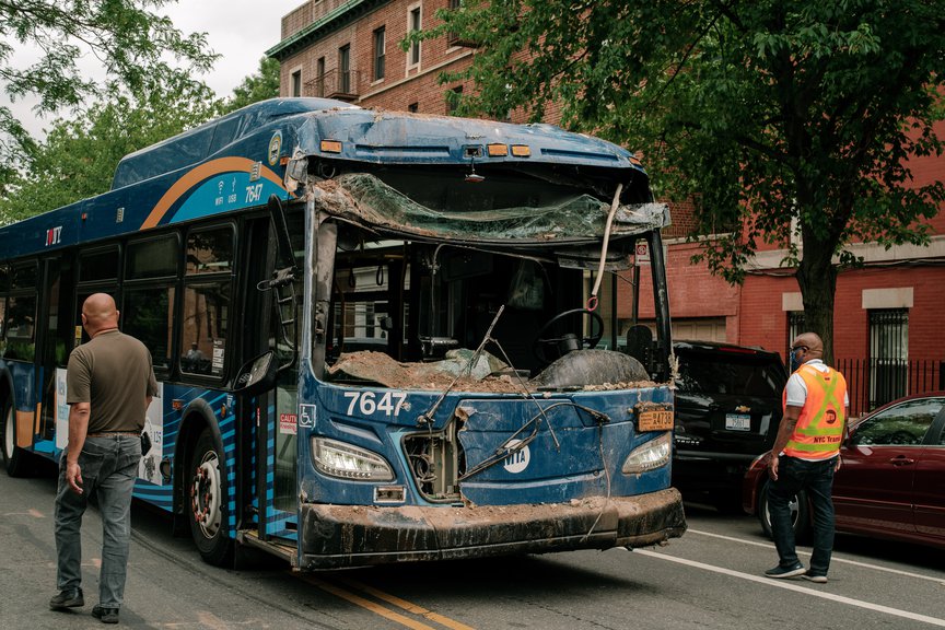 The damaged bus, with its front window shattered, and dust from the building is on the street