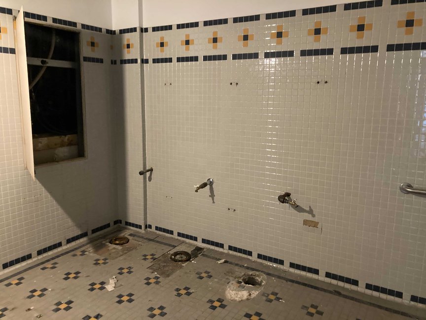 A tiled bathroom–stripped of fixtures—is seen. The tiles are white and grey, with a blue and yellow floral pattern accenting them