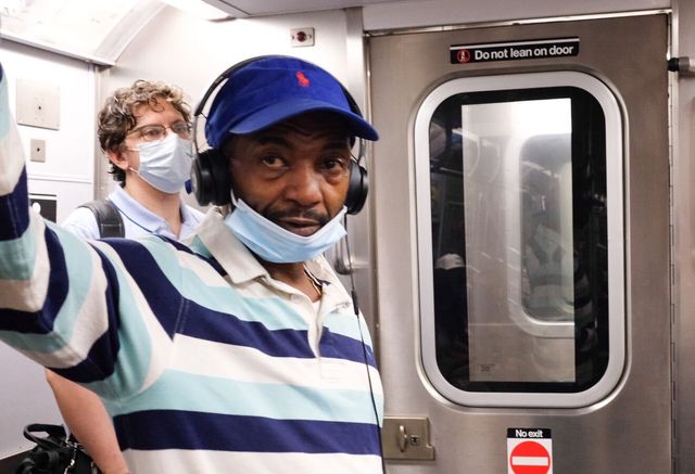 Two men ride the subway, one wearing a mask, the other with a mask tucked below his chin.